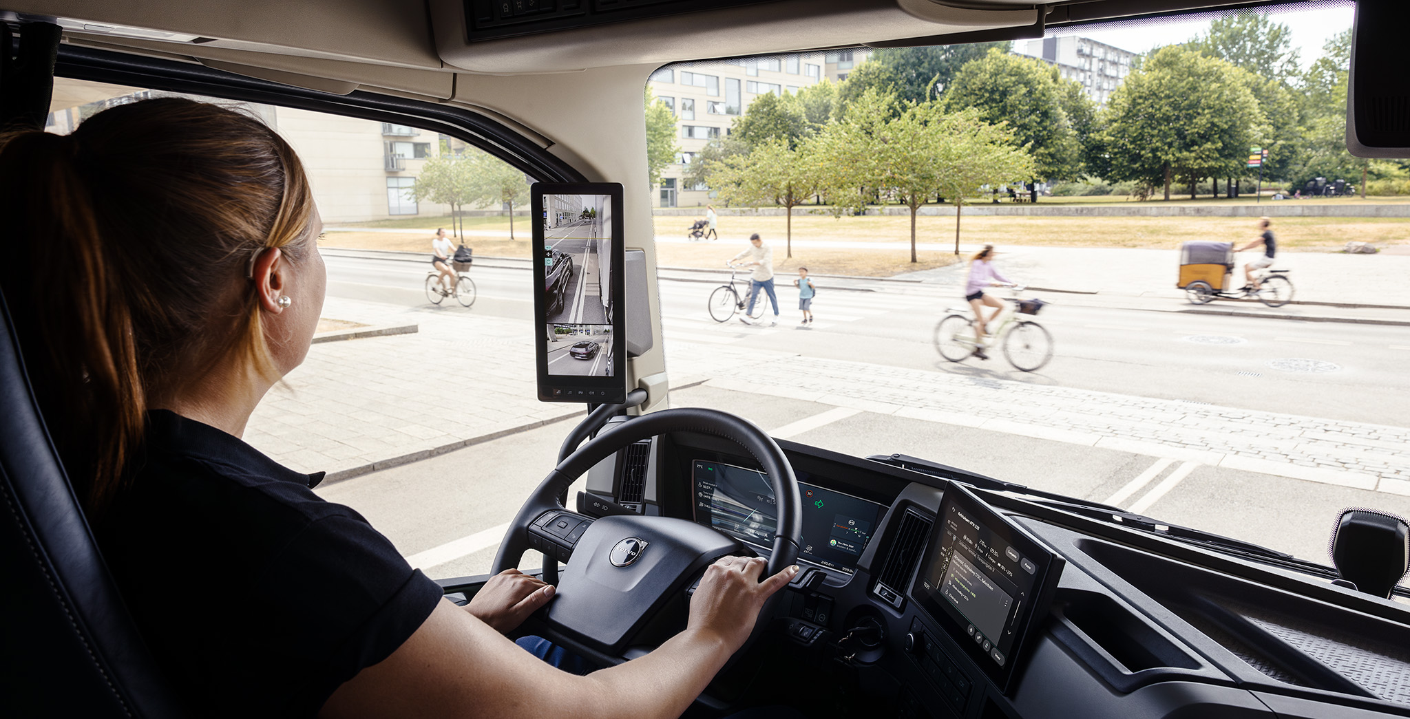 Conducteur de camion dans une cabine moderne utilisant des rétroviseurs latéraux numériques tout en naviguant dans une rue urbaine avec des cyclistes et des piétons.